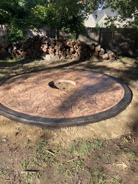 Circular stamped concrete patio with fire pit and stacked firewood in a yard.