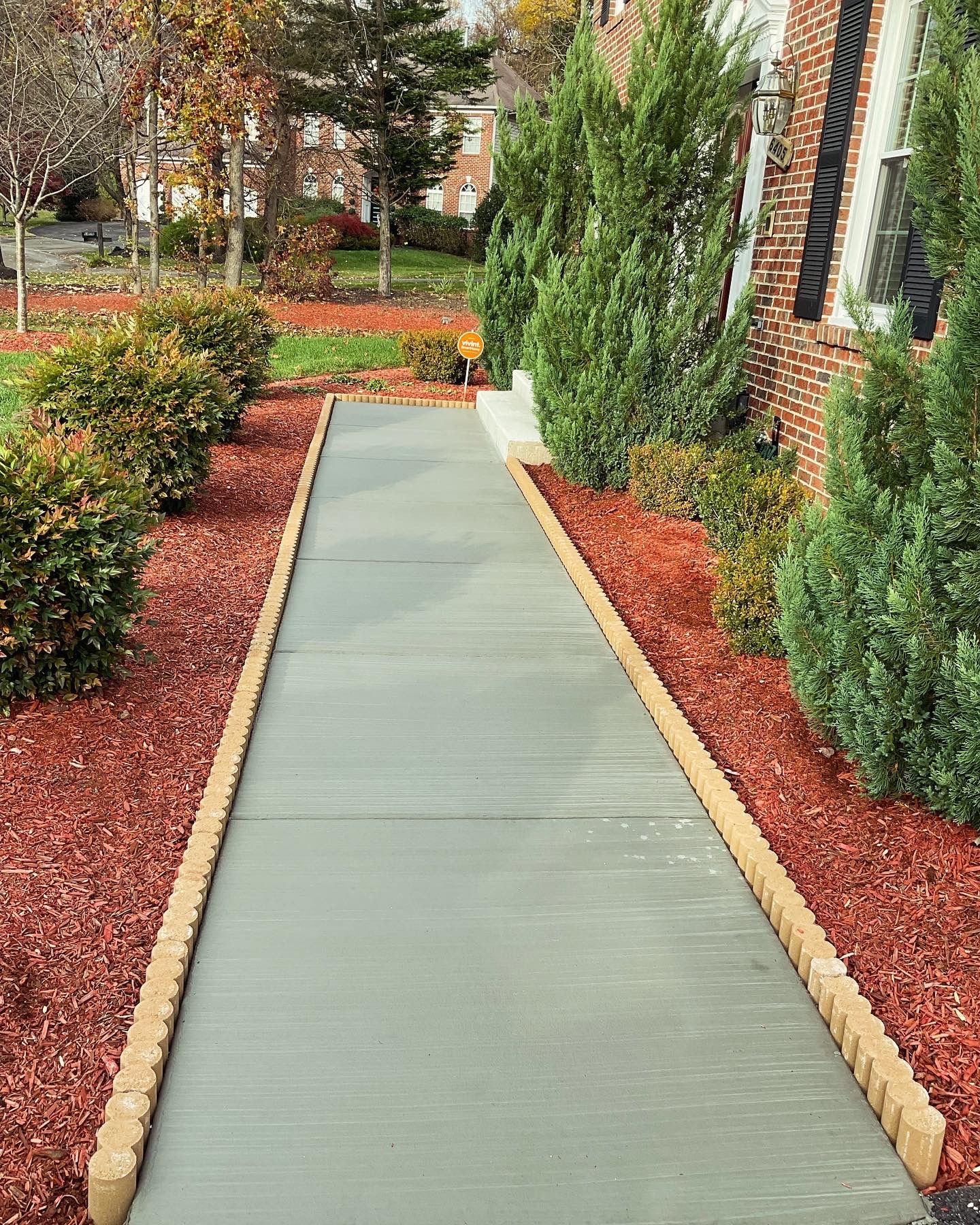 Concrete walkway bordered by red mulch and shrubs leading to a brick house.