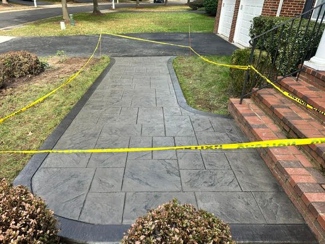 Gray patterned concrete walkway with caution tape, leading to brick steps and a driveway.