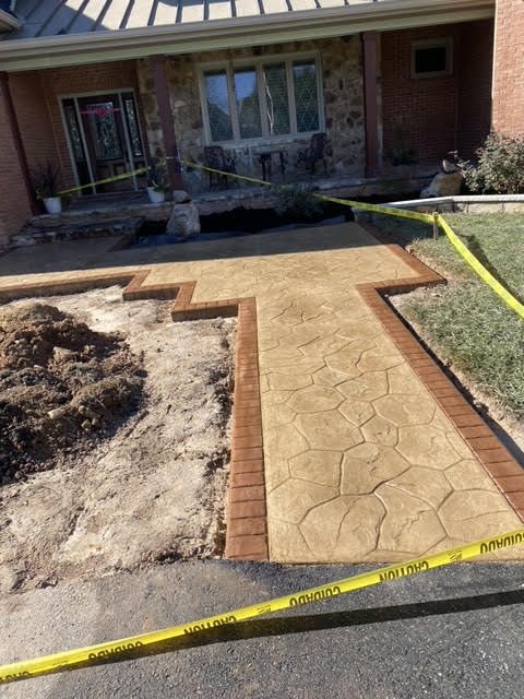 Newly constructed concrete walkway with brick-colored borders; unfinished dirt area, house in background.