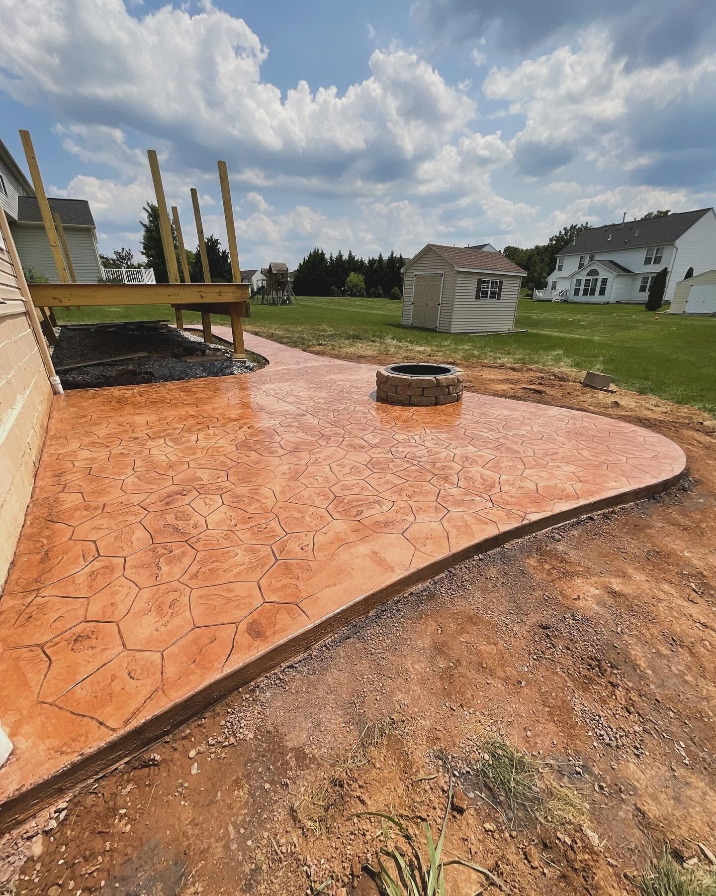Stamped concrete patio with a fire pit, next to a house and deck, under a cloudy sky.